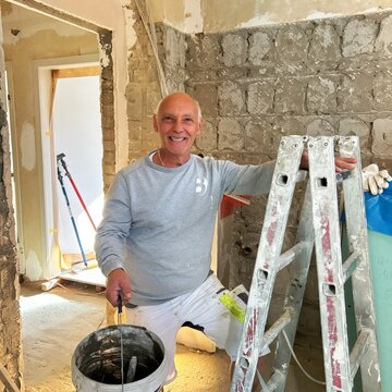Ein Handwerker lächelt freundlich, während er mit einer Kelle in einer Baustelle steht. Er hält einen Eimer und ist in Arbeitskleidung gekleidet. Im Hintergrund sind unvollendete Wände zu sehen, was auf Renovierungsarbeiten hinweist.