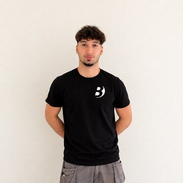 Ein junger Mann steht vor einer hellen Wand. Er trägt ein schwarzes T-Shirt mit einem weißen Logo auf der linken Brustseite. Seine Hände sind hinter dem Rücken verschränkt, und er blickt direkt in die Kamera.