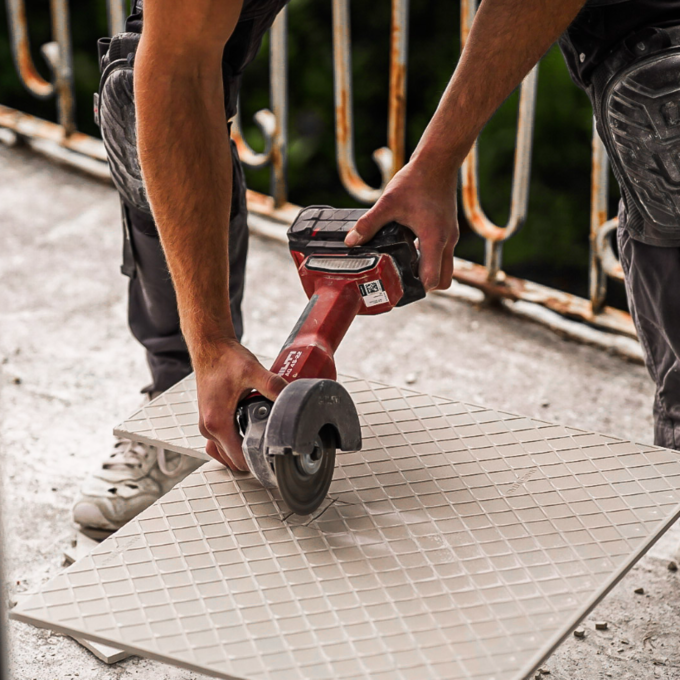 Ein Handwerker schneidet eine Fliese mit einem elektrischen Cutter. Die Fliese ist quadratisch und hat ein genopptes Muster. Der Arbeiter trägt eine Arbeitskleidung und Schutzpolster. Im Hintergrund ist ein Balkon mit einer Brüstung sichtbar.