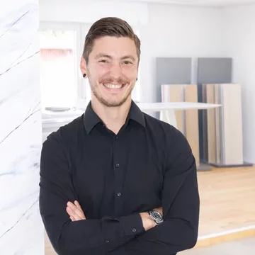 Ein Mann in einem schwarzen Hemd lächelt freundlich, während er mit überkreuzten Armen vor einem modernen Raum steht. Im Hintergrund sind verschiedene Materialien oder Möbel sichtbar. Der Raum wirkt hell und einladend.