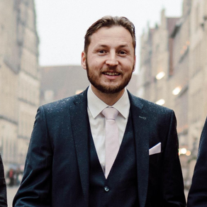 Drei Männer in eleganter Kleidung gehen gemeinsam eine Straße entlang. Der Mann in der Mitte trägt einen dunklen Anzug mit einem hellen Hemd und einer Krawatte. Er lächelt freundlich, während die anderen beiden leicht im Hintergrund stehen. Historische Gebäude sind im Hintergrund sichtbar.