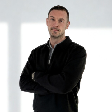 Ein Mann steht mit verschränkten Armen in einem hellen, minimalistisch gestalteten Raum. Er trägt einen schwarzen Pullover und schaut direkt in die Kamera. Die Wände sind weiß und es gibt sanfte Lichtreflexionen im Hintergrund.