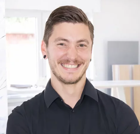 Ein junger Mann mit kurzen, dunklen Haaren lächelt freundlich in die Kamera. Er trägt ein schwarzes Hemd und hat Ohrstecker. Im Hintergrund sind verschwommene Möbel und Fenster zu sehen, die einen hellen, modernen Raum zeigen.