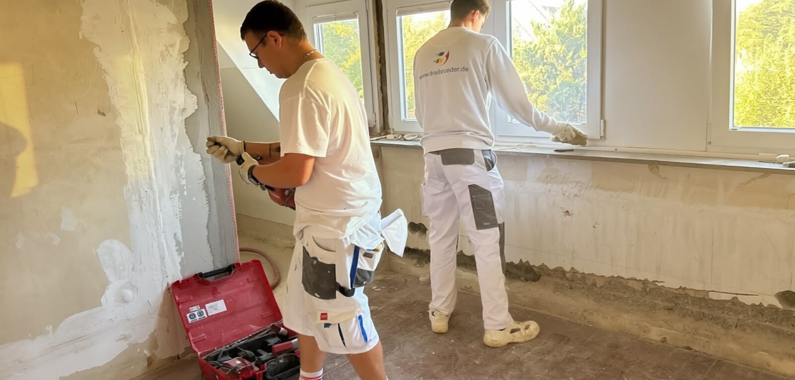 Zwei Handwerker arbeiten in einem Raum. Der erste, links, trägt eine weiße T-Shirt und eine Werkzeugkiste, während er an einer Wand arbeitet. Der zweite, rechts, ist dabei, eine Fensterbank zu bearbeiten. Es gibt mehrere Fenster und unvollendete Wandflächen im Hintergrund.