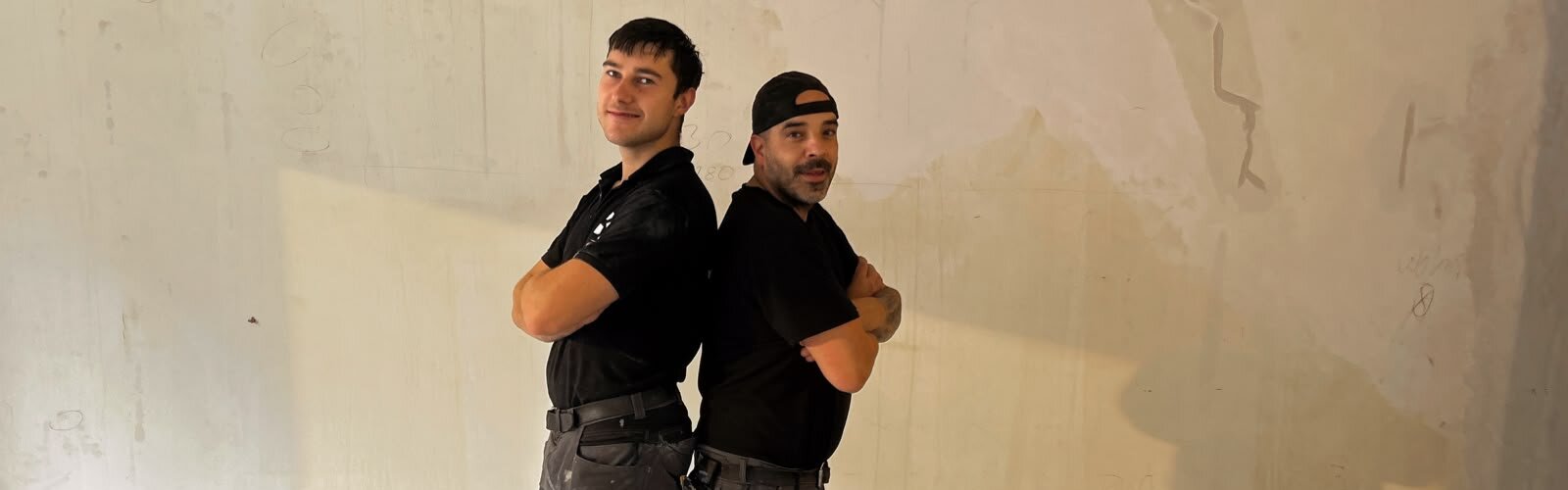 Zwei junge Handwerker stehen Rücken an Rücken in einem leeren Raum. Beide tragen schwarze Arbeitskleidung und spezielle Knieschützer. Der Raum hat einen hellen, neutralen Hintergrund mit unvollendeten Wänden. Der Ausdruck der jungen Männer ist entspannt und selbstbewusst.