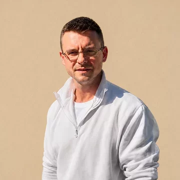 Ein Mann mit kurzen, braunen Haaren steht vor einem hellen Hintergrund. Er trägt eine weiße Jacke mit Reißverschluss und eine weiße T-Shirt. Der Mann schaut direkt in die Kamera und hat eine neutrale Miene.
