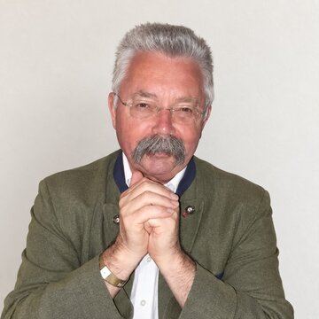 Ein älterer Mann mit grauen Haaren und einem markanten Schnurrbart trägt einen grünen Blazer. Er sitzt mit gefalteten Händen vor einem hellen Hintergrund und blickt freundlich in die Kamera. Seine Haltung wirkt nachdenklich und einladend.