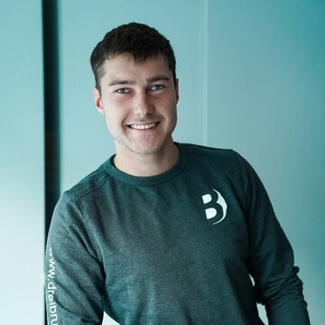 Ein junger Mann lächelt, während er an einem Tisch lehnt. Er trägt ein graues Sweatshirt mit einem Logo. Im Vordergrund sind einige gestapelte Objekte zu sehen, deren Details nicht erkennbar sind. Der Hintergrund ist hell und minimalistisch.