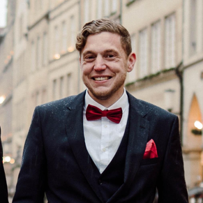 Ein Mann in einem eleganten Anzug mit roter Fliege und Einstecktuch steht lächelnd auf einer regnerischen Straßenszene. Im Hintergrund sind historische Gebäude und Fahrräder sichtbar.