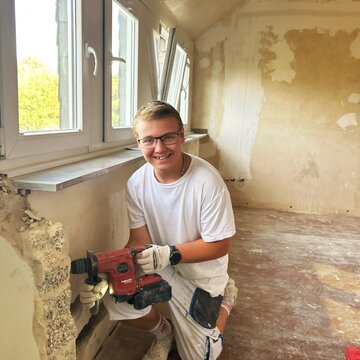 Ein lächelnder junger Mann sitzt auf dem Boden eines renovierungsbedürftigen Raums und hält ein Elektrowerkzeug. Er trägt ein weißes T-Shirt und Handschuhe. Im Hintergrund sind Fenster und unvollendete Wände sichtbar.