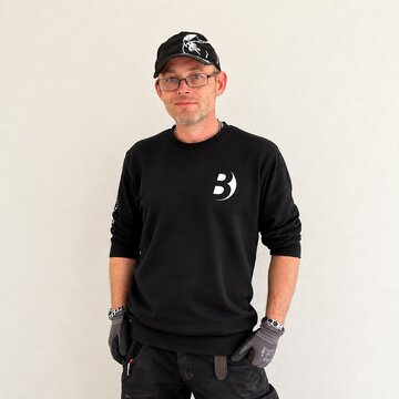 Ein Mann steht vor einer hellen Wand. Er trägt ein schwarzes Sweatshirt mit einem weißen Logo und eine schwarze Cap. Der Mann hat eine Brille und lächelt leicht. Seine Hände sind teilweise in Handschuhen, und er hat kurze, gepflegte Haare.