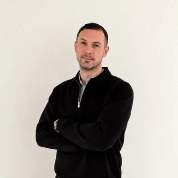 Ein Mann steht mit verschränkten Armen vor einem neutralen, hellen Hintergrund. Er trägt einen schwarzen Pullover und blickt direkt in die Kamera. Sein Ausdruck wirkt selbstbewusst und freundlich.