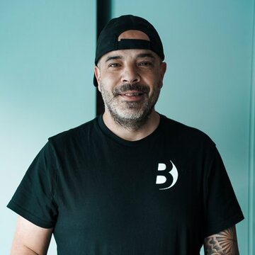 Ein Mann mit einem kurzen Bart und einem schwarzen T-Shirt steht an einem Tisch mit bunten Unterlagen. Er trägt eine Baseballcap und lächelt freundlich in die Kamera. Der Hintergrund ist hell und minimalistisch.